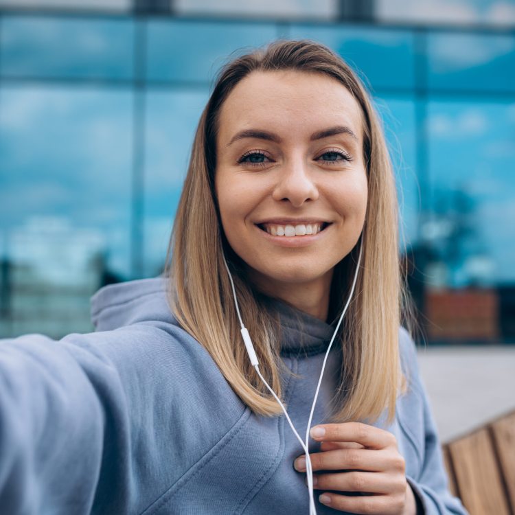 Front view of girl in earphones taking selfie outdoors. Pretty young blonde sitting on bench, holding smartphone by one hand, looking at camera. Concept of modern lifestyle.